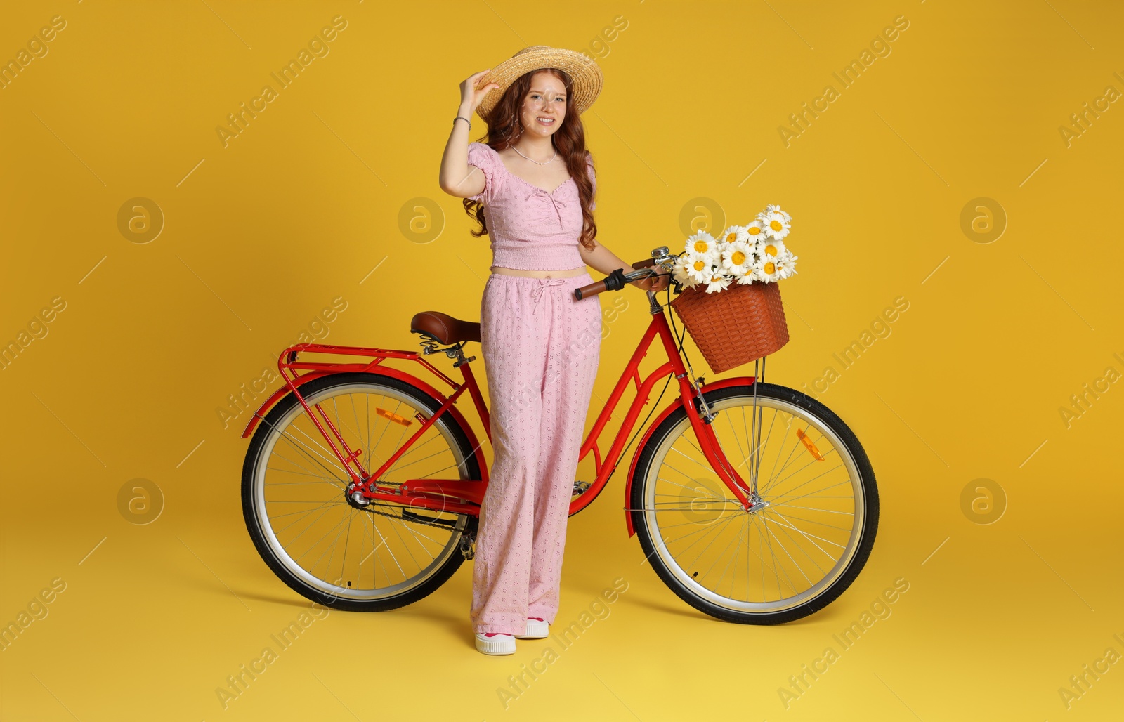 Smiling teenage girl in hat and bicycle with basket of flowers against yellow background Photo of Smiling teenage girl in hat and bicycle with basket of flowers against yellow background