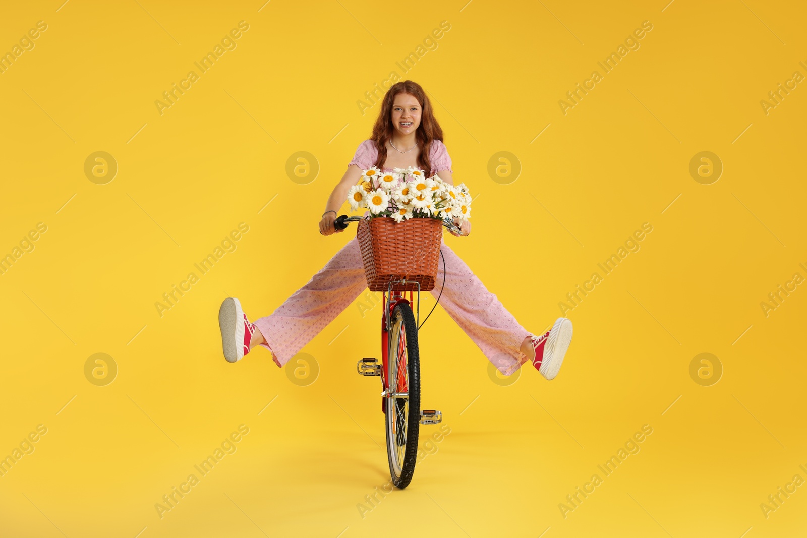 Smiling teenage girl riding bicycle with basket of flowers against yellow background Photo of Smiling teenage girl riding bicycle with basket of flowers against yellow background