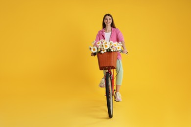 Photo of Smiling woman riding bicycle with basket of flowers against yellow background. Space for text