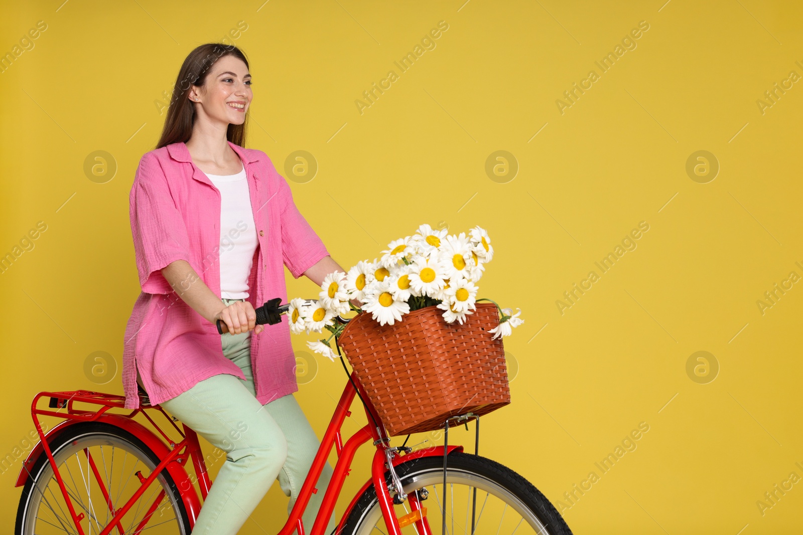 Smiling woman riding bicycle with basket of flowers against yellow background. Space for text Photo of Smiling woman riding bicycle with basket of flowers against yellow background. Space for text
