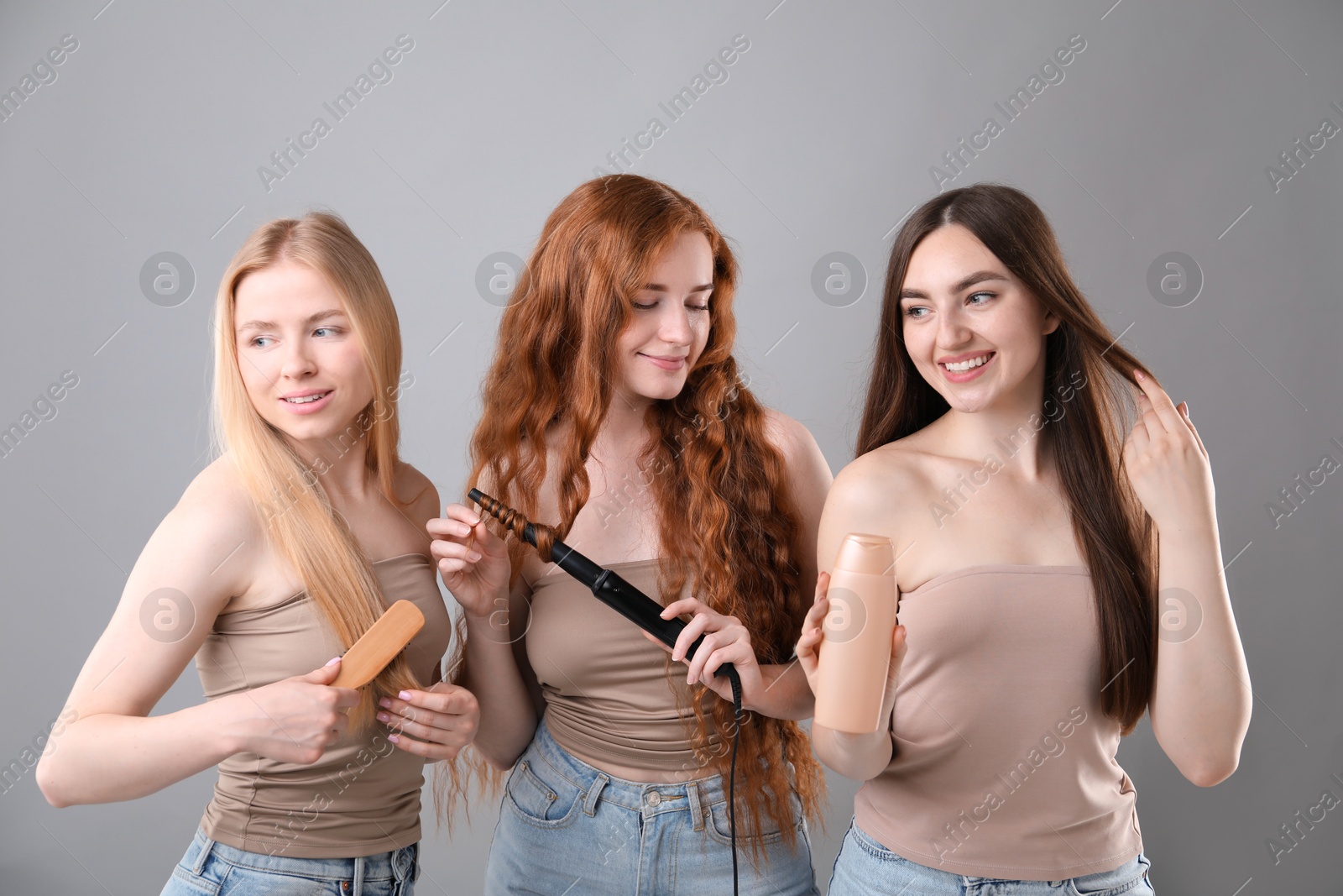Beautiful women with different hair colors holding curling iron, shampoo and brush on grey background Photo of Beautiful women with different hair colors holding curling iron, shampoo and brush on grey background