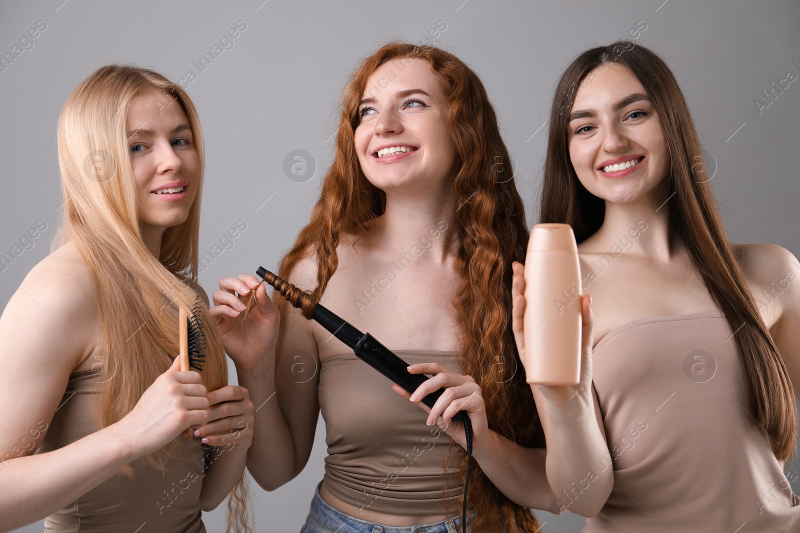 Photo of Beautiful women with different hair colors holding curling iron, shampoo and brush on grey background