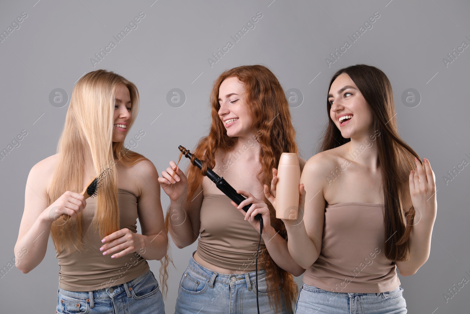 Beautiful women with different hair colors holding curling iron, shampoo and brush on grey background Photo of Beautiful women with different hair colors holding curling iron, shampoo and brush on grey background