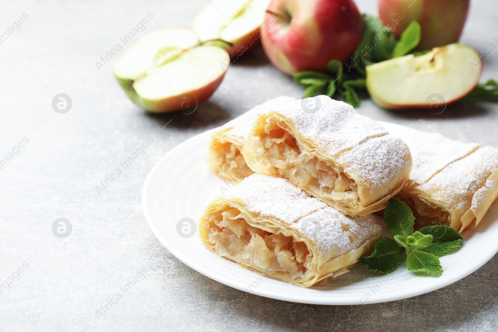 Photo of Pieces of tasty apple strudel with powdered sugar and mint on light grey table, closeup