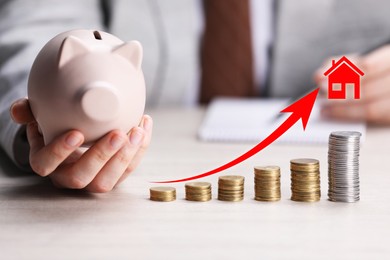 Image of Increase in real estate value. Arrow and house icon above stacked coins. Man holding piggy bank at table, closeup