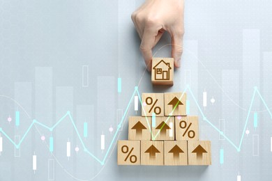 Image of Increase in real estate value. Woman putting cube with house icon to others on grey background, top view. Percent signs and arrows on wooden cubes