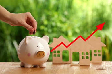 Image of Increase in real estate value. Woman putting coin into piggy bank outdoors, closeup. House models and arrow