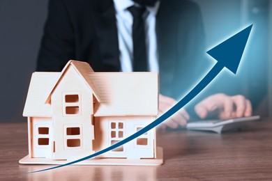 Image of Increase in real estate value. House model and arrow. Man using calculator at table, selective focus