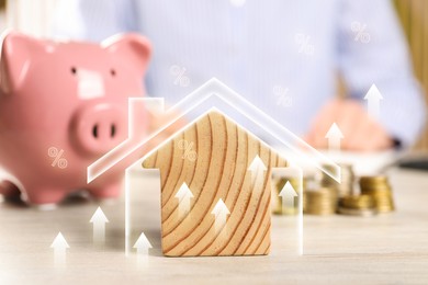 Image of Increase in real estate value. House model, piggy bank and coins on wooden table. Growing price represented by arrows and percent signs