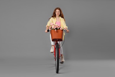 Photo of Beautiful teenage girl riding bicycle with basket of flowers against grey background