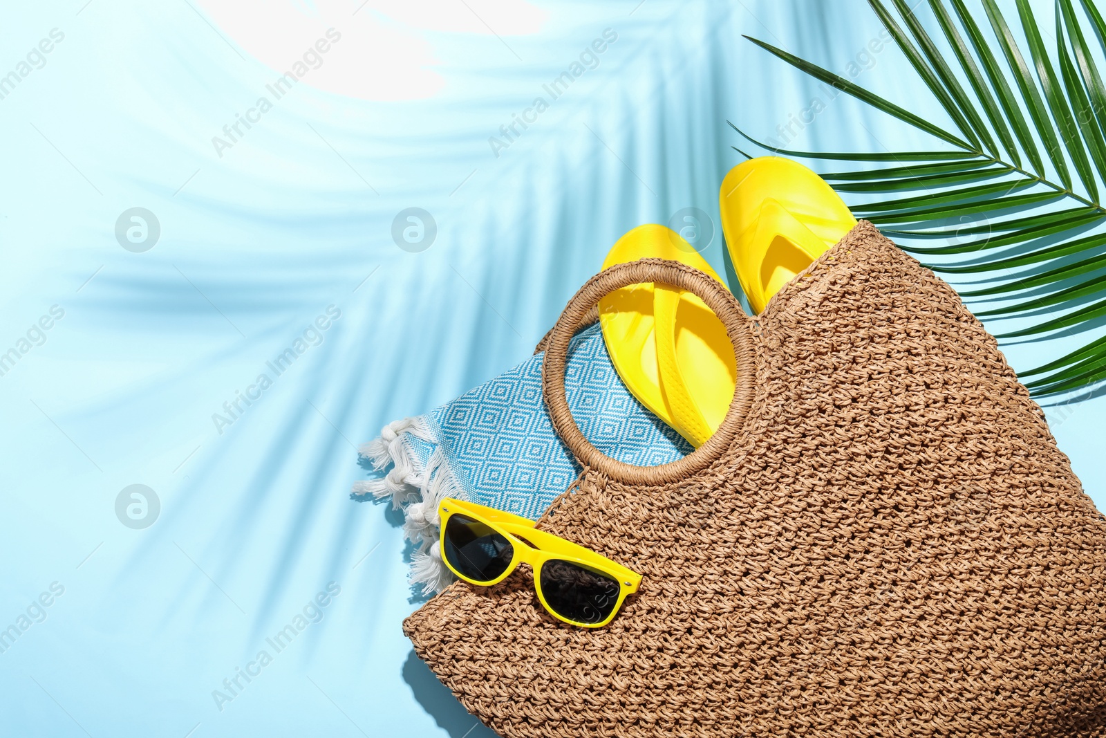Wicker bag with beach towel, sunglasses, flip flops and palm leaf on light blue background, flat lay. Space for text Photo of Wicker bag with beach towel, sunglasses, flip flops and palm leaf on light blue background, flat lay. Space for text