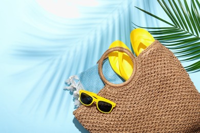 Photo of Wicker bag with beach towel, sunglasses, flip flops and palm leaf on light blue background, flat lay. Space for text