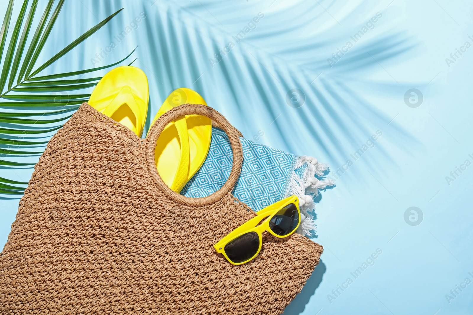 Wicker bag with beach towel, sunglasses, flip flops and palm leaf on light blue background, flat lay. Space for text Photo of Wicker bag with beach towel, sunglasses, flip flops and palm leaf on light blue background, flat lay. Space for text