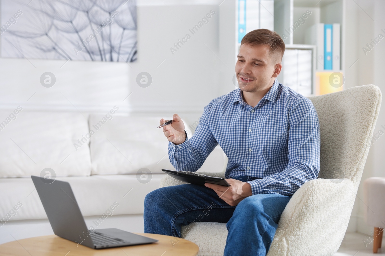 Smiling psychologist with clipboard during online session by laptop in office Photo of Smiling psychologist with clipboard during online session by laptop in office