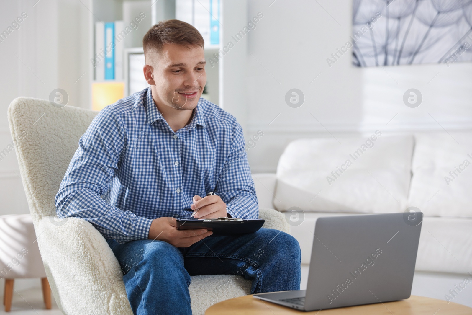 Smiling psychologist with clipboard during online session by laptop in office Photo of Smiling psychologist with clipboard during online session by laptop in office