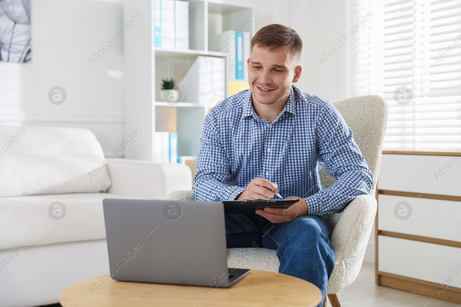 Smiling psychologist with clipboard during online session by laptop in office Photo of Smiling psychologist with clipboard during online session by laptop in office