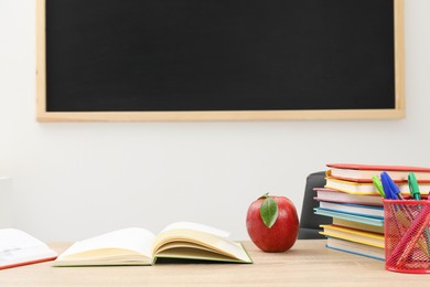 Photo of Different stationery and red apple on wooden table near blackboard indoors