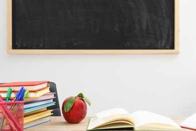 Photo of Different stationery and red apple on wooden table near blackboard indoors, closeup