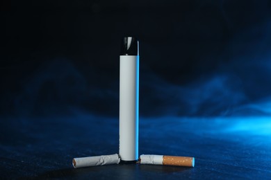 Photo of Electronic smoking device and broken cigarette on black table in blue light