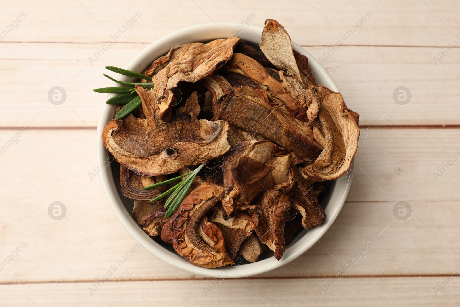 Pieces of dried mushrooms and rosemary in bowl on light wooden table, top view Photo of Pieces of dried mushrooms and rosemary in bowl on light wooden table, top view