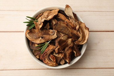 Photo of Pieces of dried mushrooms and rosemary in bowl on light wooden table, top view