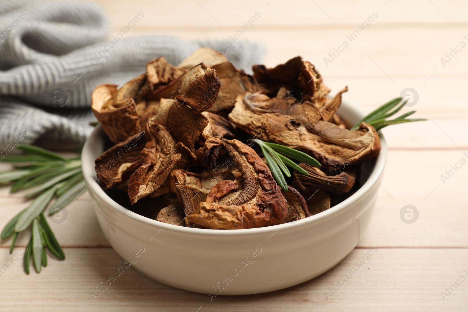 Photo of Pieces of dried mushrooms and rosemary in bowl on light wooden table, closeup