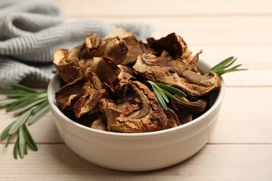 Photo of Pieces of dried mushrooms and rosemary in bowl on light wooden table, closeup