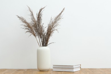 Photo of Dried reeds in vase and books on wooden table near white wall indoors