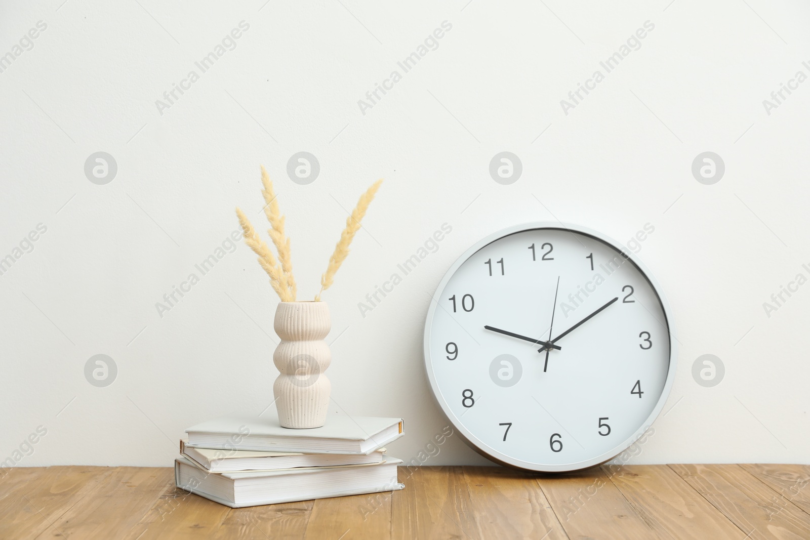 Dried spikes, books and round clock on wooden table near white wall indoors Photo of Dried spikes, books and round clock on wooden table near white wall indoors