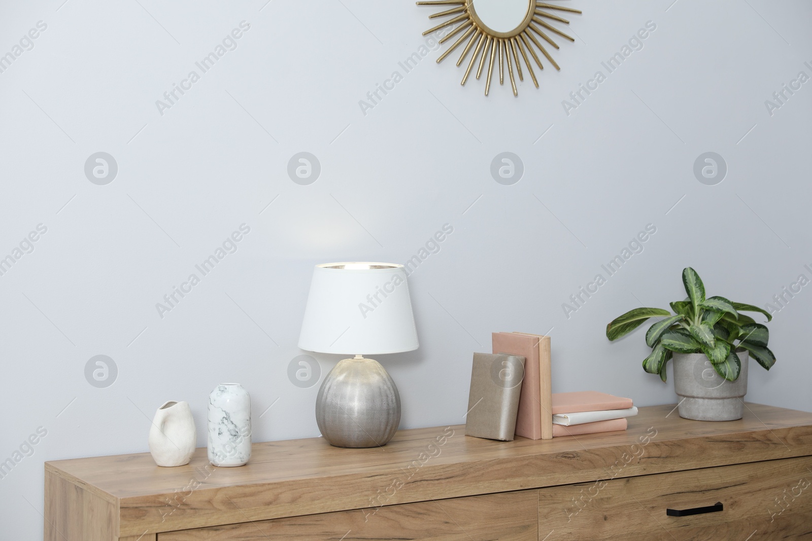 Lamp, books, vases and green houseplant on wooden cabinet near white wall indoors Photo of Lamp, books, vases and green houseplant on wooden cabinet near white wall indoors