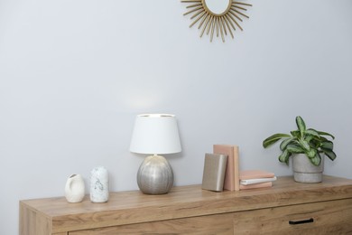 Photo of Lamp, books, vases and green houseplant on wooden cabinet near white wall indoors