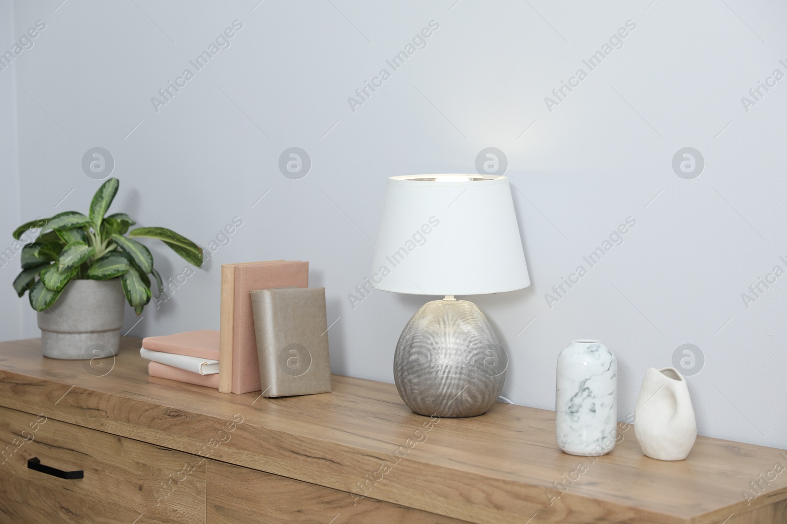 Lamp, books, vases and green houseplant on wooden cabinet near white wall indoors Photo of Lamp, books, vases and green houseplant on wooden cabinet near white wall indoors
