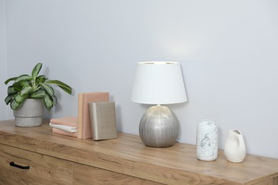 Photo of Lamp, books, vases and green houseplant on wooden cabinet near white wall indoors
