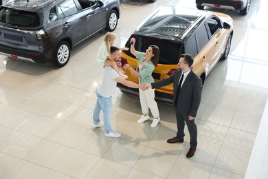 Photo of Salesman shaking hands with happy family near new car in salon