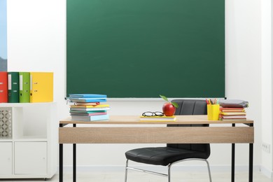 Photo of Different stationery, glasses and red apple on wooden table near blackboard indoors