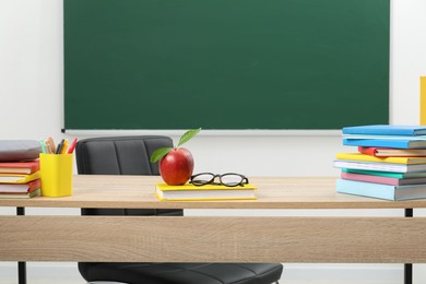 Photo of Different stationery, glasses and red apple on wooden table near blackboard indoors