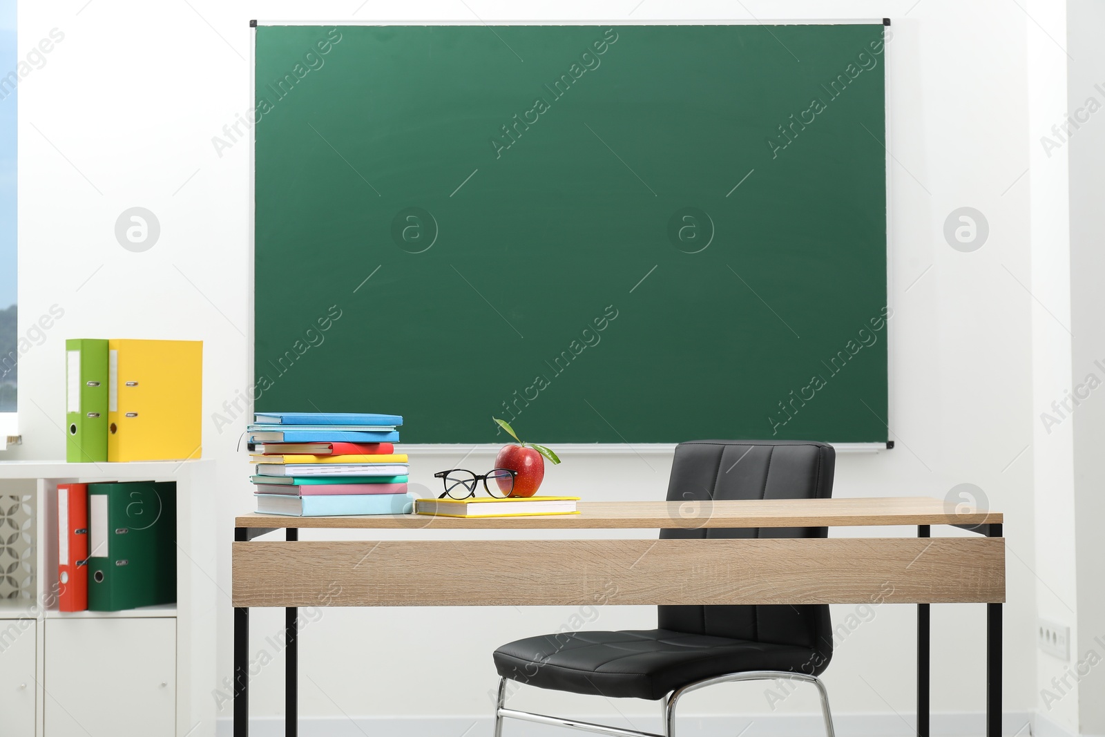 Different books, glasses and red apple on wooden table near blackboard indoors Photo of Different books, glasses and red apple on wooden table near blackboard indoors