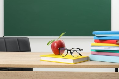 Photo of Different books, glasses and red apple on wooden table near blackboard indoors, space for text