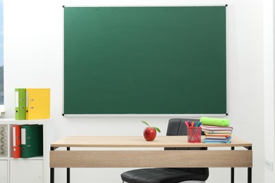 Photo of Different stationery and red apple on wooden table near blackboard indoors