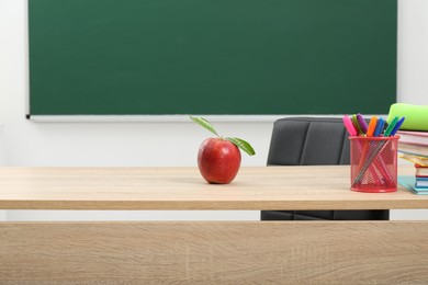 Photo of Different stationery and red apple on wooden table near blackboard indoors