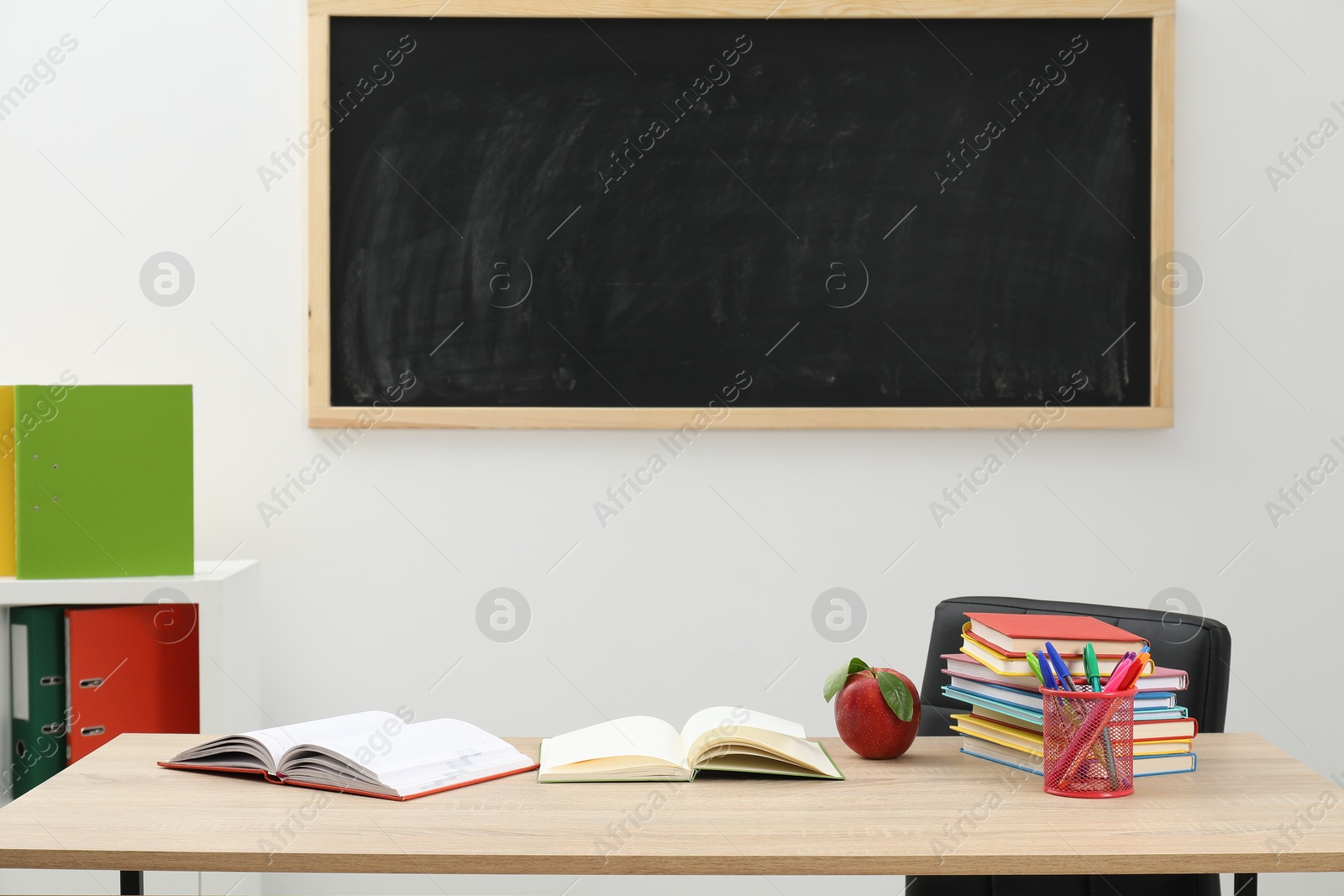 Different stationery and red apple on wooden table near blackboard indoors Photo of Different stationery and red apple on wooden table near blackboard indoors