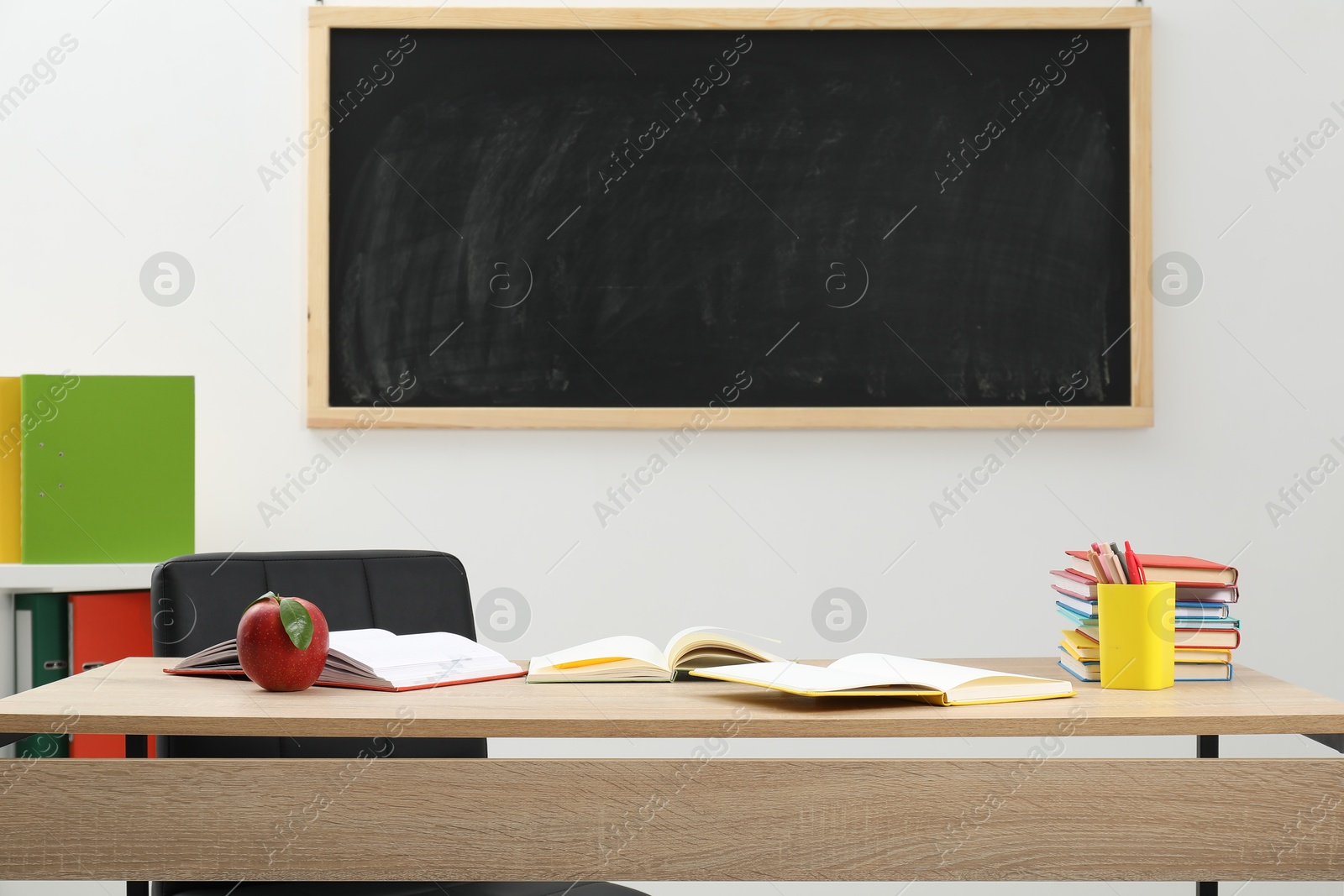 Different stationery and red apple on wooden table near blackboard indoors Photo of Different stationery and red apple on wooden table near blackboard indoors