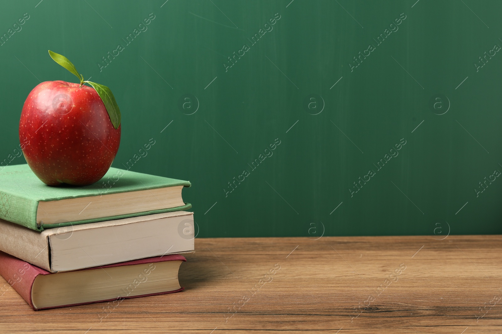 Different books and red apple on wooden table near blackboard, closeup. Space for text Photo of Different books and red apple on wooden table near blackboard, closeup. Space for text
