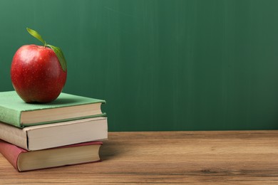 Photo of Different books and red apple on wooden table near blackboard, closeup. Space for text