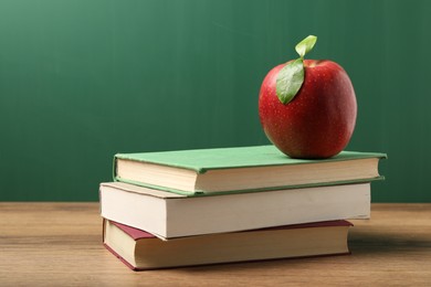 Photo of Different books and red apple on wooden table near blackboard, closeup