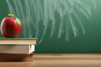 Photo of Different books and red apple on wooden table near blackboard, closeup. Space for text