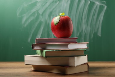 Photo of Different books and red apple on wooden table near blackboard, closeup