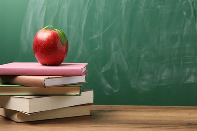 Photo of Different books and red apple on wooden table near blackboard, closeup. Space for text