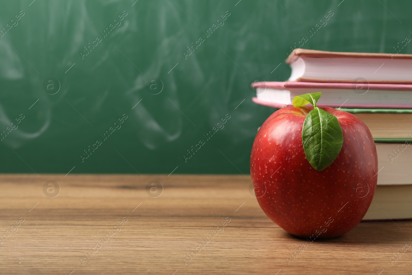 Different books and red apple on wooden table near blackboard, closeup. Space for text Photo of Different books and red apple on wooden table near blackboard, closeup. Space for text
