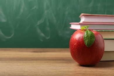 Photo of Different books and red apple on wooden table near blackboard, closeup. Space for text
